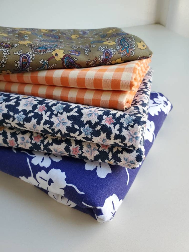 May include: A stack of four different fabric swatches. The top swatch is a green paisley print, the second is a orange and white gingham, the third is a black and white floral print, and the bottom is a dark blue floral print.