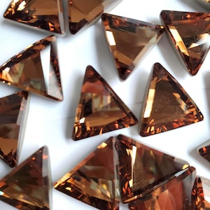 May include: A collection of small, triangular, faceted glass rhinestones in a warm brown color. The rhinestones are scattered on a white surface.
