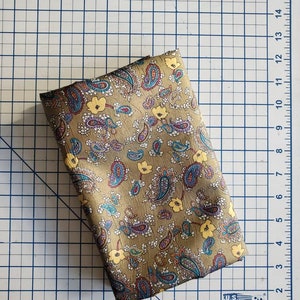 May include: A folded piece of fabric with a paisley pattern in shades of brown, blue, yellow, and white. The fabric is on a gridded surface.