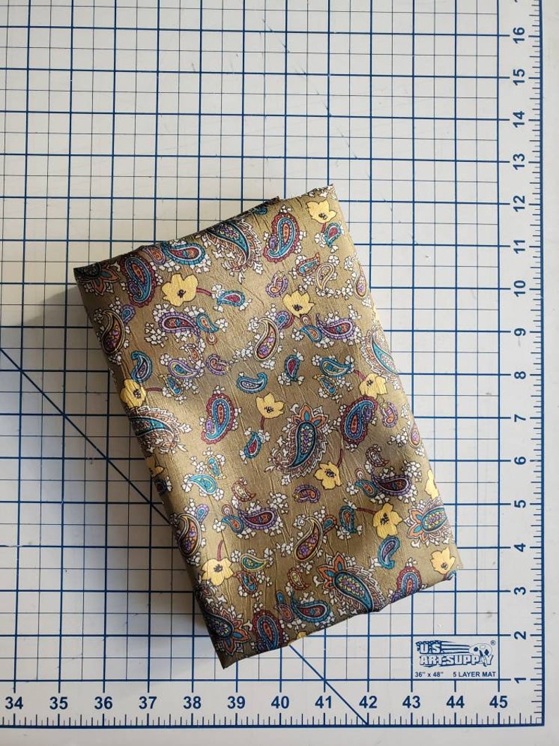 May include: A folded piece of fabric with a paisley pattern in shades of brown, blue, yellow, and white. The fabric is on a gridded surface.