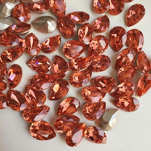 May include: A collection of pear-shaped, orange-colored rhinestones. The rhinestones are scattered on a white surface.