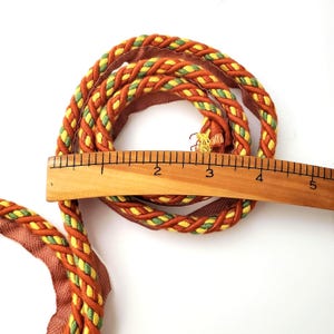 Puede incluir: Ribete trenzado en tonos naranja, amarillo y verde, con reverso marrón. Una regla de madera se coloca sobre el ribete, mostrando medidas en centímetros. El ribete está enrollado sobre una superficie blanca.