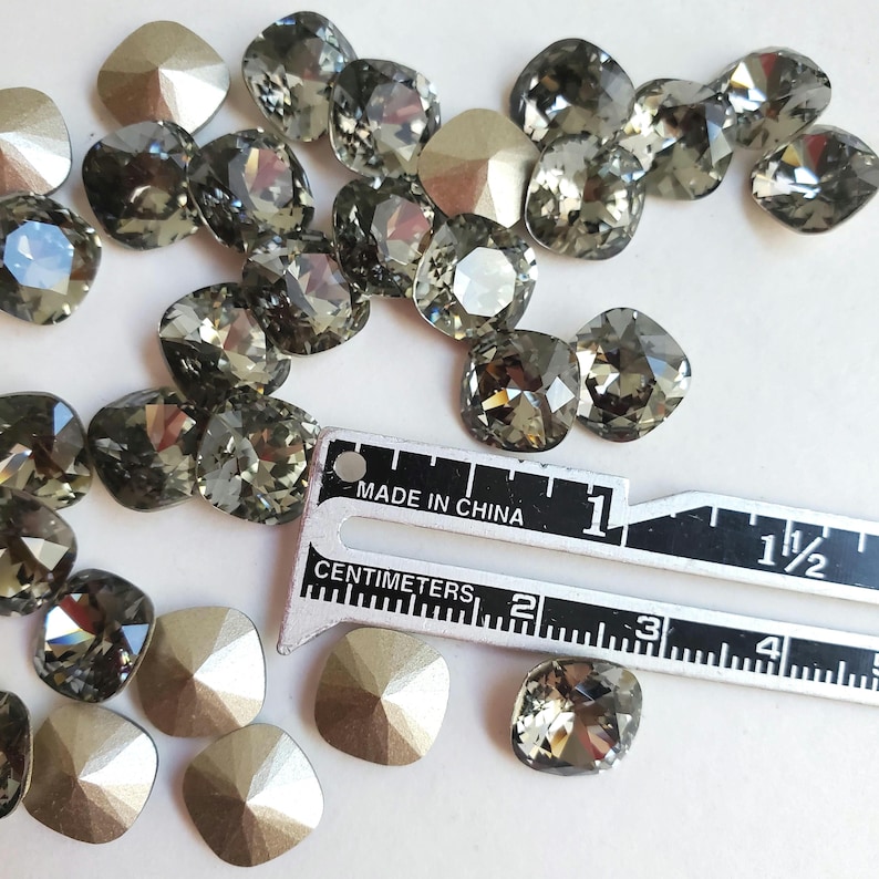 Pu&ograve; includere: Primo piano di un gruppo di strass quadrati grigi e dorati. Gli strass sono sparsi su una superficie bianca accanto a un righello graduato in pollici e centimetri. "Made in China" &egrave; stampato sul righello.