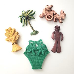 Könnte beinhalten: Eine Sammlung von fünf Kunststoffanstecknadeln mit einer gelben Weizengarbe, einer grünen Palme, einer rosafarbenen Figur mit Esel und Karren, einer braunen stehenden Figur und einem grünen Korb mit Blumen. Diese Anstecknadeln haben geformte Details.
