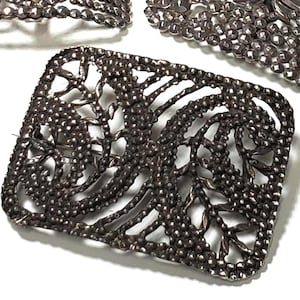 May include: Close-up of a rectangular, ornate silver-toned brooch with a detailed openwork design. The brooch features a textured surface with a pattern of small, raised dots and leaf-like shapes. Other matching jewelry pieces are visible.