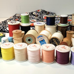 Pode incluir: Uma coleção de carretéis de linha em várias cores, incluindo verde, amarelo, rosa, laranja, roxo e azul. Os carretéis estão dispostos em uma fileira sobre uma superfície branca. Alguns dos carretéis têm etiquetas com a marca "Clark's" e o texto "Mercerized 300 Yds."