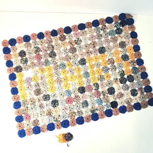 Op de afbeelding: Een rechthoekige, handgemaakte quilt met een patchwork-ontwerp van kleine, cirkelvormige stukjes stof in verschillende kleuren, waaronder geel, roze, blauw en bruin. De quilt is afgezet met blauwe cirkels. Een kleine, bijpassende speld ligt naast de quilt.