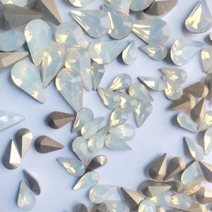 May include: A large assortment of iridescent white teardrop-shaped rhinestones. The rhinestones are scattered on a white surface.