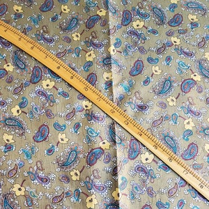 May include: A close-up of a fabric with a paisley pattern. The fabric is a light brown color with a subtle sheen. The paisley pattern is in shades of blue, purple, and yellow. A wooden ruler is placed diagonally across the fabric.