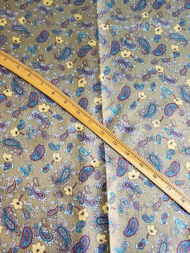 May include: A close-up of a fabric with a paisley pattern. The fabric is a light brown color with a subtle sheen. The paisley pattern is in shades of blue, purple, and yellow. A wooden ruler is placed diagonally across the fabric.