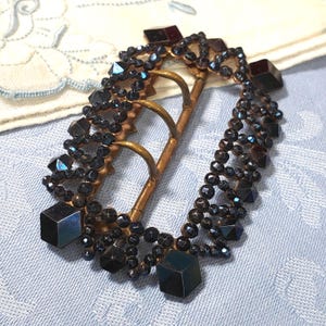 May include: An antique oval buckle with a brass frame and three loops. The buckle is adorned with dark, iridescent beads and square-cut accents. The beads are arranged in a decorative pattern around the frame.