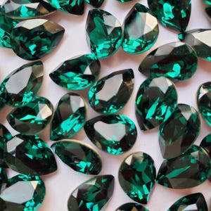 Puede incluir: Primer plano de un grupo de piedras de imitación de forma de pera de color verde esmeralda. Las piedras de imitación están esparcidas sobre una superficie blanca.