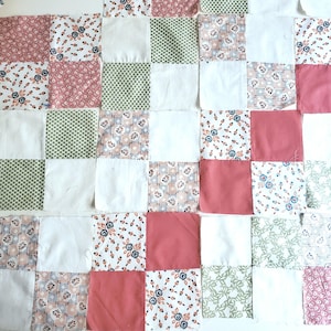 May include: A patchwork quilt in progress, featuring a checkerboard pattern of squares. The squares are in various colors and patterns, including white, dusty rose, sage green, and floral prints. The quilt is laid out on a white surface.