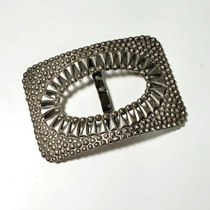 May include: Silver-toned rectangular belt buckle with an oval center. The buckle is heavily embellished with small, round studs around the perimeter and triangular accents around the oval opening. A metal bar with two holes is attached to the back.