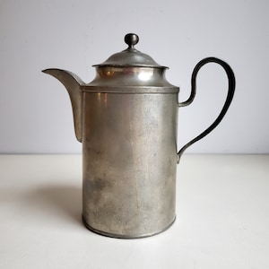 May include: A silver pitcher with a black handle and a silver lid. The pitcher has a rounded body and a spout on the side.