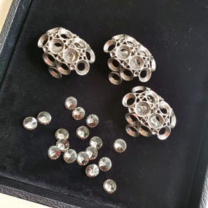 Könnte beinhalten: Drei silberne Broschen aus Metall mit einem runden Design und klaren Strasssteinen. Die Broschen sind in einer Reihe auf einer schwarzen Oberfläche angeordnet. Es gibt auch mehrere klare Strasssteine, die um die Broschen herum verstreut sind.