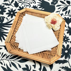 Può includere: Un vassoio rettangolare in rattan intrecciato contiene una pila di tovaglioli di lino bianco con bordi cuciti. Un grande fiore color crema con un centro rosso è appoggiato sul vassoio. Il vassoio poggia su un tessuto a fantasia floreale bianco e nero.