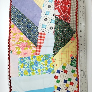 May include: A patchwork quilt with a red, white, and blue color scheme. The quilt is made of various fabrics with different patterns, including floral, geometric, and plaid. The quilt is edged with a red scalloped trim. A ruler is visible in the image.