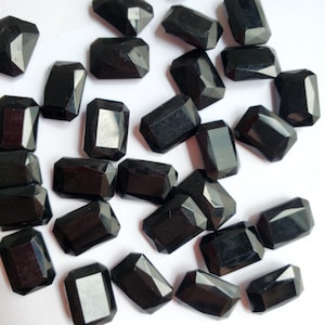 May include: A collection of faceted, rectangular black gemstones. The gemstones have a polished, reflective surface and are scattered across a white background. The shapes are similar to emerald cuts.