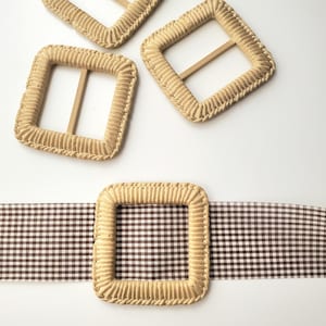 May include: Three square belt buckles wrapped in light beige cord, featuring rounded corners and a central horizontal bar. One buckle is threaded onto a brown and white gingham fabric belt, while two additional buckles are positioned above it.
