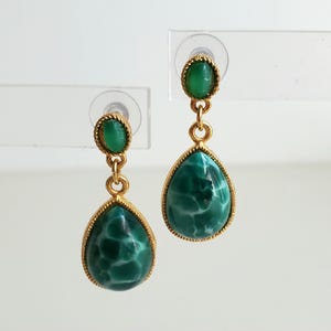 May include: A pair of gold-toned drop earrings. Each earring features a small, oval-shaped green stone at the top, connected by a gold chain to a larger, teardrop-shaped green stone with a marbled pattern. The earrings are set against a white background.
