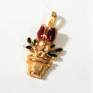 May include: A gold-toned pendant featuring a potted plant design. The pendant showcases a textured pot, golden stems, green leaves, and two red flower buds. The pendant has a loop at the top for a chain.