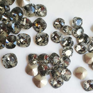 Pu&ograve; includere: Una collezione di strass quadrati di colore grigio scuro, alcuni con finitura sfaccettata, sparsi su una superficie bianca.