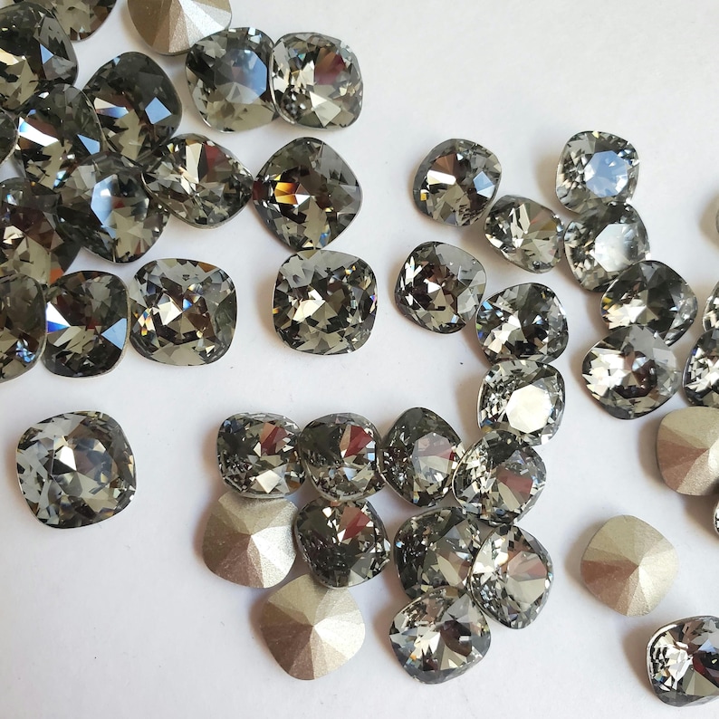 Pu&ograve; includere: Una collezione di strass quadrati di colore grigio scuro, alcuni con finitura sfaccettata, sparsi su una superficie bianca.
