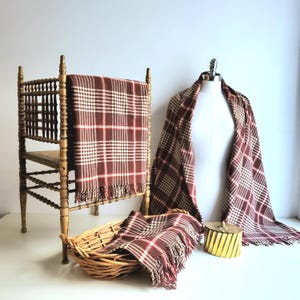 May include: A vintage-style display featuring a brown and cream plaid scarf draped over a wooden chair, a mannequin, and in a wicker basket. The scarf has a fringed edge. A small gold-colored container with a lid is also present.