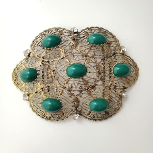 May include: Vintage gold-tone brooch with a floral filigree design. The brooch features seven oval, emerald-green cabochon stones and small clear rhinestones. The brooch is an ornate, statement piece.
