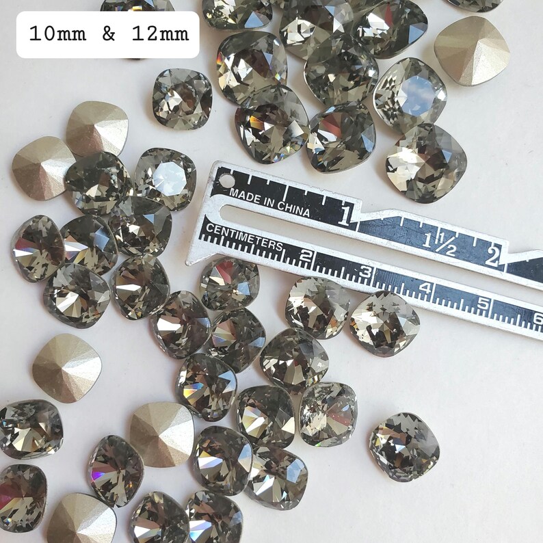 Pu&ograve; includere: Una collezione di strass quadrati di colore grigio scuro da 10 mm e 12 mm. Gli strass sono sparsi su una superficie bianca con un righello per riferimento alle dimensioni.  MADE IN CHINA &egrave; stampato sul righello.
