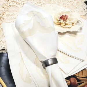 May include: White linen napkins with a light beige leaf design, one folded and held by a silver napkin ring. A matching placemat and a decorative flower on a white dish are also visible. The overall theme is elegant dining.
