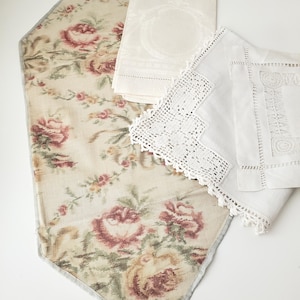 Può includere: Una collezione di tre pezzi tessili che include un lungo runner da tavola con sfondo beige e un motivo floreale di rose rosse e verdi, un panno color crema piegato con un sottile disegno intrecciato e un panno quadrato bianco con bordo in pizzo.