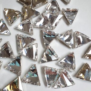 May include: A collection of clear, triangular, faceted rhinestones. The rhinestones have a silver-toned finish and are scattered on a white surface.