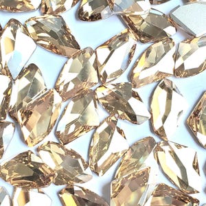 Puede incluir: Una colección de cristales de vidrio facetados de color champán claro con una forma geométrica de escudo irregular. Cada cristal presenta múltiples superficies pulidas que reflejan la luz, creando una apariencia brillante y translúcida en todo el conjunto.