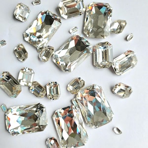 Puede incluir: Una colección de gemas de vidrio rectangulares transparentes y facetadas, esparcidas sobre una superficie blanca. Las gemas son de varios tamaños y tienen un efecto arcoíris iridiscente.