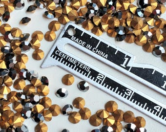 May include: A close-up of a large quantity of gold and silver rhinestones. The rhinestones are scattered on a white surface. A ruler with the text "Made in China" and "Centimeters" is placed on the surface.