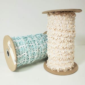May include: Two spools of decorative trim. One spool is a light blue and white woven design. The other spool is a cream-colored trim with a textured, looped design. Both spools are on cardboard spools.