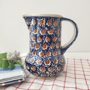 Może przedstawiać: Ceramiczny dzban z niebiesko-brązowym wzorem w kropki. Dzbanek ma białą obręcz i uchwyt. Dzbanek stoi na czerwono-białym obrusie w kratę.