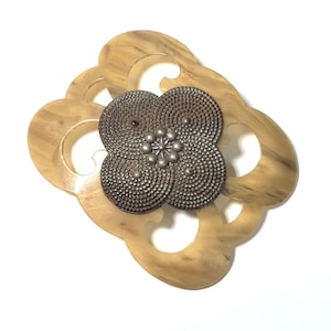 以下が含まれることがあります： ヴィンテージのブローチ。薄茶色の渦巻き模様の透かし彫りのフレーム。中央には、質感のある濃い色の金属で作られた四弁の花のデザインがあり、中央には銀色のビーズのクラスターがあります。ブローチは白い背景に配置されています。