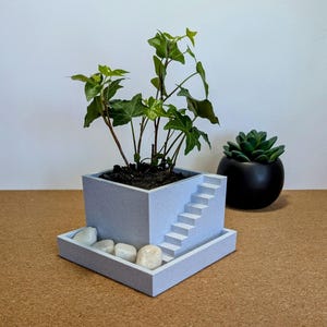Peut inclure: Un pot carré bleu clair avec un design d'escalier miniature, contenant une plante verte. Le pot repose sur un plateau assorti avec des pierres blanches. Un pot rond noir avec une succulente verte est à droite.