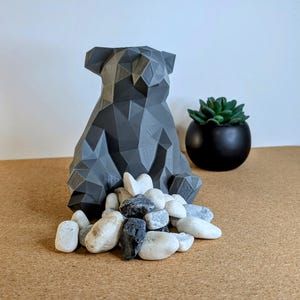 May include: A gray geometric bear figurine sits on a cork surface with white and black pebbles around it. A small black pot with a green succulent plant is in the background.