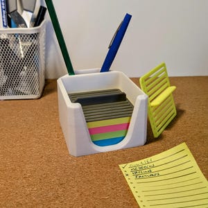 Puede incluir: Organizador de escritorio blanco impreso en 3D con una ranura para bolígrafos y un compartimento para notas adhesivas. El organizador tiene un porta notas adhesivas gris, rosa, amarillo, verde y azul. Un bloc de notas amarillo con una lista de verificación que dice "GUINEA, MEDICIN, VACCINS, FACTLURS" está en el escritorio.