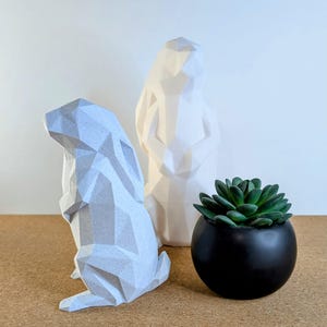 May include: Two white geometric animal sculptures, one of a rabbit and one of a person, with a small black pot containing a green succulent plant.