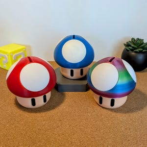 Puede incluir: Tres huchas con forma de hongo en rojo, azul y arcoíris. Cada una tiene una parte superior blanca con una ranura para monedas y una base beige con dos puntos negros. Un bloque amarillo con un signo de interrogación está en el fondo.