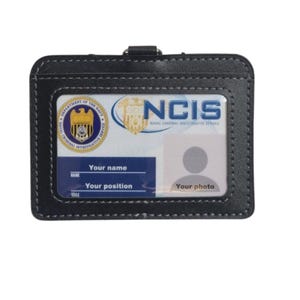 May include: Black leather badge holder with a clear plastic insert for an ID card. The holder has a metal clip for attaching to a belt or lanyard. The ID card insert has the text "NCIS NAVAL CRIMINAL INVESTIGATIVE SERVICE" and a space for a photo.