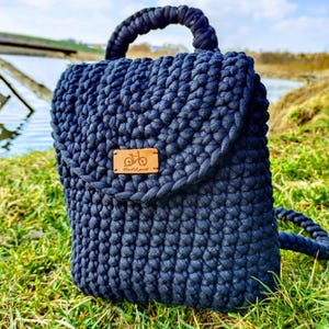 May include: A dark blue crocheted backpack with a flap closure and a brown leather patch with a bicycle logo and the text "handmade" on it.