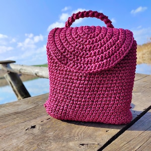 May include: A pink crocheted bag with a rounded flap and a top handle. The bag is sitting on a wooden surface.