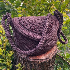 May include: A handmade, crochet shoulder bag in a deep plum color. The bag features a rounded flap and a braided strap. The bag is sitting on a tree stump, with green foliage in the background.