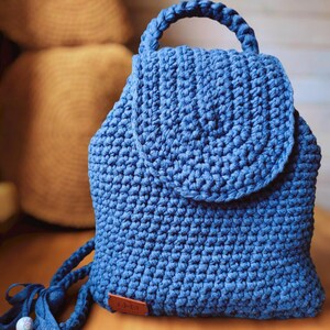 May include: A blue crochet backpack featuring a thick top handle, a rounded flap closure, and a textured stitch pattern. The bag includes a small brown rectangular tag at the bottom corner and braided straps with a light-colored bead detail.